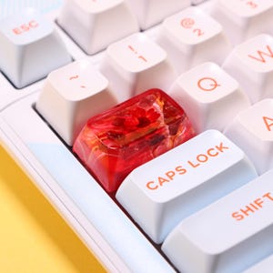 May include: Close-up of a white keyboard with a custom red keycap. The keycap is transparent with a floral design. The words "CAPS LOCK" are visible on a white key.