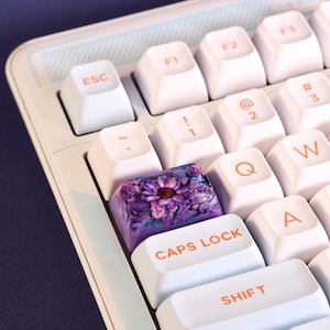 May include: Close-up of a computer keyboard with white keys and orange lettering. The "CAPS LOCK" key features a clear resin design with a purple flower. The keyboard has a light blue and white striped design.