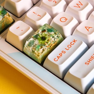 May include: Close-up of a computer keyboard with custom keycaps. The keycaps are white with orange lettering. One keycap is a clear resin with green and yellow floral inclusions. The keyboard has a light blue and yellow accent.