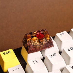 May include: A close-up of a custom keyboard keycap with a red, gold, and brown resin design. The keycap is sitting on a white keyboard.