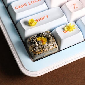 Peut inclure: Gros plan sur un clavier avec des touches blanches et un bord bleu clair. Les touches portent des lettres oranges, dont "CAPS LOCK" et "SHIFT". Une touche est une touche personnalisée en résine avec des paillettes dorées et une fleur jaune.