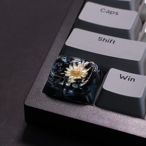 Peut inclure: Une touche de clavier personnalisée avec une petite fleur séchée encapsulée dans de la résine transparente. La touche a un effet marbré bleu foncé et blanc, et est placée sur un clavier gris. Les touches adjacentes sont étiquetées "Caps", "Shift" et "Win".