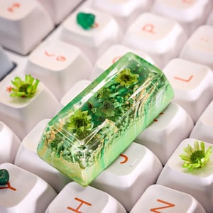 May include: A green and white resin keycap with embedded dried flowers and green glitter. The keycap is sitting on a white keyboard.