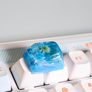 Peut inclure: Un capuchon de touche en résine bleu et blanc avec une fleur pressée à l'intérieur. Le capuchon de touche est posé sur un clavier blanc.