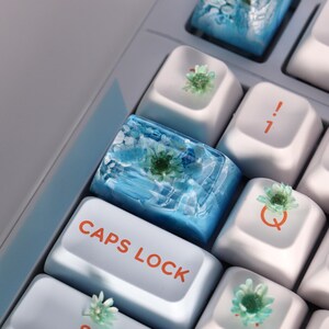 May include: Close-up of a keyboard with custom keycaps. The keycaps are white with floral accents and a blue resin keycap with a flower. The "CAPS LOCK" key is visible in the image.