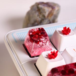 Peut inclure: Gros plan sur un capuchon de touche en résine rose et blanche avec une fleur rouge intégrée. Le capuchon de touche est sur un clavier blanc avec d'autres capuchons de touche présentant des fleurs rouges.