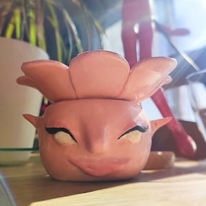 Puede incluir: Un macetero de cerámica rosa con forma de cara, con ojos grandes y una amplia sonrisa. El macetero tiene una parte superior con forma de flor.