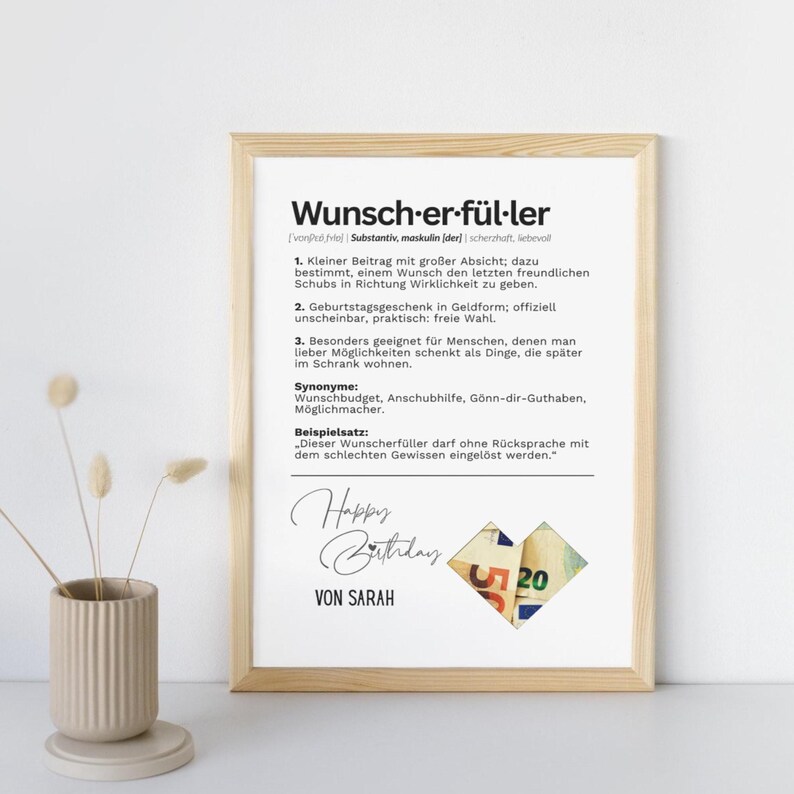 May include: A framed print with the German word "Wunscherf&uuml;ller" and its definition. The print includes the text "Happy Birthday" and "Von Sarah", with a collage of money. The frame is a light brown color.
