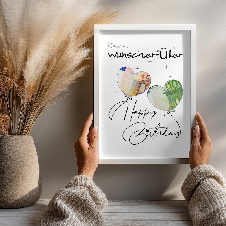 K&ouml;nnte beinhalten: Ein wei&szlig;er Bilderrahmen mit einem Druck, der den Text "kleiner Wunscherf&uuml;ller" und "Happy Birthday" in schwarzer Schrift zeigt. Zwei herzf&ouml;rmige Ballon-Illustrationen enthalten Bilder eines 50- und 100-Euro-Scheins, verziert mit kleinen Sternen.