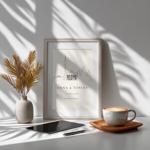 K&ouml;nnte beinhalten: Gerahmter Kunstdruck mit einer minimalistischen Strichzeichnung einer Stadtlandschaft, einschlie&szlig;lich eines Turms und Geb&auml;uden. Der Druck zeigt die Namen "ANNA & TOBIAS" und das Datum "14. JULI 2026". Eine Vase mit getrockneten Pflanzen, ein Tablet und eine Kaffeetasse sind ebenfalls sichtbar.
