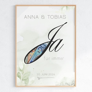 May include: A framed print featuring the text ANNA & TOBIAS, the word Ja, and the phrase für immer. The letter J contains a cutout revealing Euro banknotes. Below is the date 20. JUNI 2026 and a small line drawing of a ring. The background has green leaves.