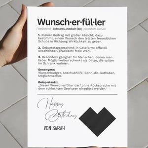 May include: A white card with German text, held by hands. The card features the words "Wunsch-er-f&uuml;ller" and "Happy Birthday" in script, along with a black heart-shaped design. The card is a birthday gift.