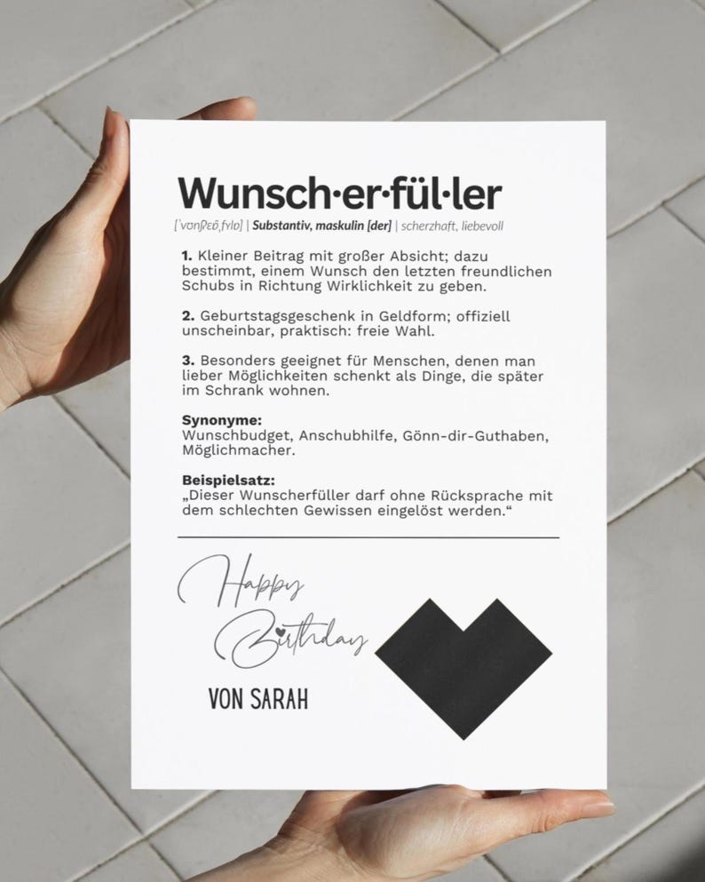 May include: A white card with German text, held by hands. The card features the words "Wunsch-er-f&uuml;ller" and "Happy Birthday" in script, along with a black heart-shaped design. The card is a birthday gift.