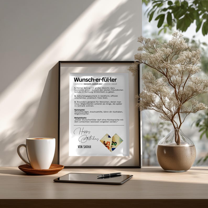 May include: A framed print with the German word "Wunsch-er-f&uuml;ller" and text. The print includes the words "Happy Birthday" and "Von Sarah". A cup of coffee, a tablet, and a vase with branches are on the table.