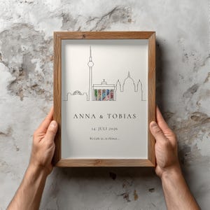 K&ouml;nnte beinhalten: Ein gerahmter Druck mit einer minimalistischen Strichzeichnung einer Stadtlandschaft, einschlie&szlig;lich eines hohen Turms und Kuppelbauten. Der Druck tr&auml;gt die Namen "ANNA & TOBIAS" und das Datum "14. JULI 2026". Der Rahmen ist aus hellem Holz gefertigt.