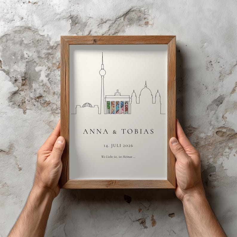K&ouml;nnte beinhalten: Ein gerahmter Druck mit einer minimalistischen Strichzeichnung einer Stadtlandschaft, einschlie&szlig;lich eines hohen Turms und Kuppelbauten. Der Druck tr&auml;gt die Namen "ANNA & TOBIAS" und das Datum "14. JULI 2026". Der Rahmen ist aus hellem Holz gefertigt.