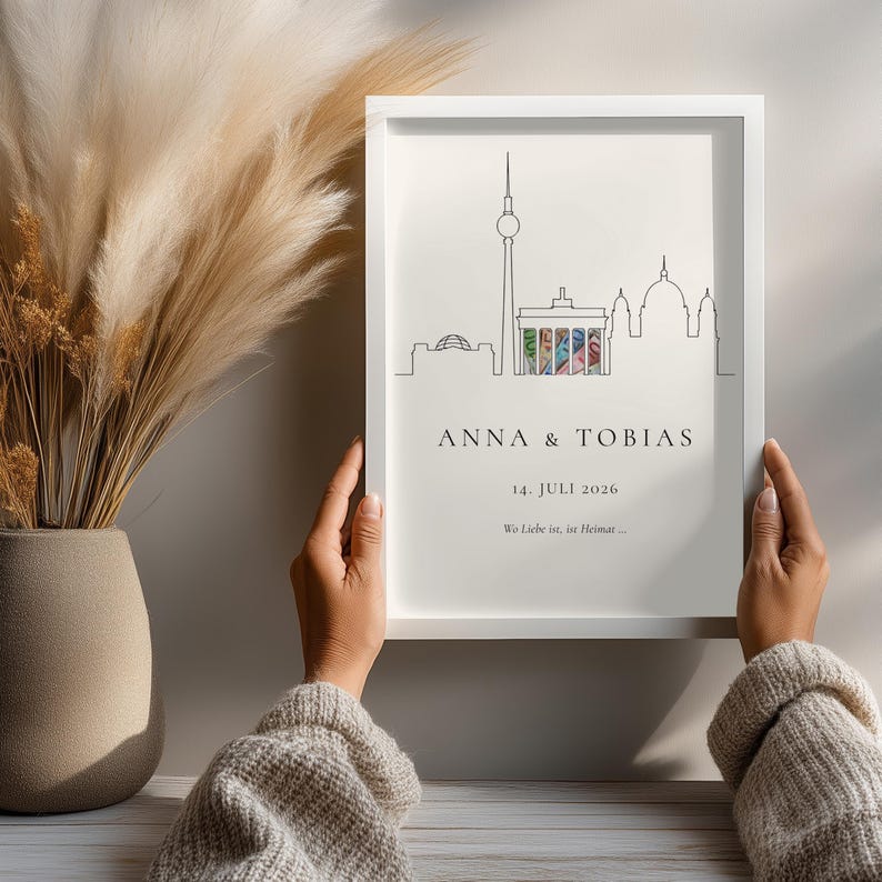 K&ouml;nnte beinhalten: Ein gerahmter Druck mit einer minimalistischen Strichzeichnung der Berliner Skyline, einschlie&szlig;lich Fernsehturm und Brandenburger Tor. Der Druck enth&auml;lt die Namen "ANNA & TOBIAS" und das Datum "14. JULI 2026".