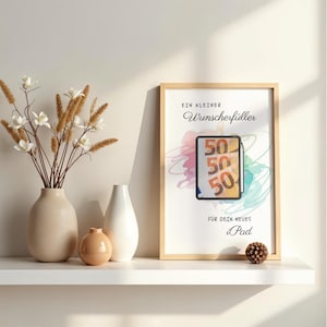 May include: A framed print featuring the German text "EIN KLEINER Wunscherf&uuml;ller F&Uuml;R DEIN neues iPad" with a central graphic of a tablet displaying fifty-euro banknotes against a colorful watercolor background, presented in a light wood frame.