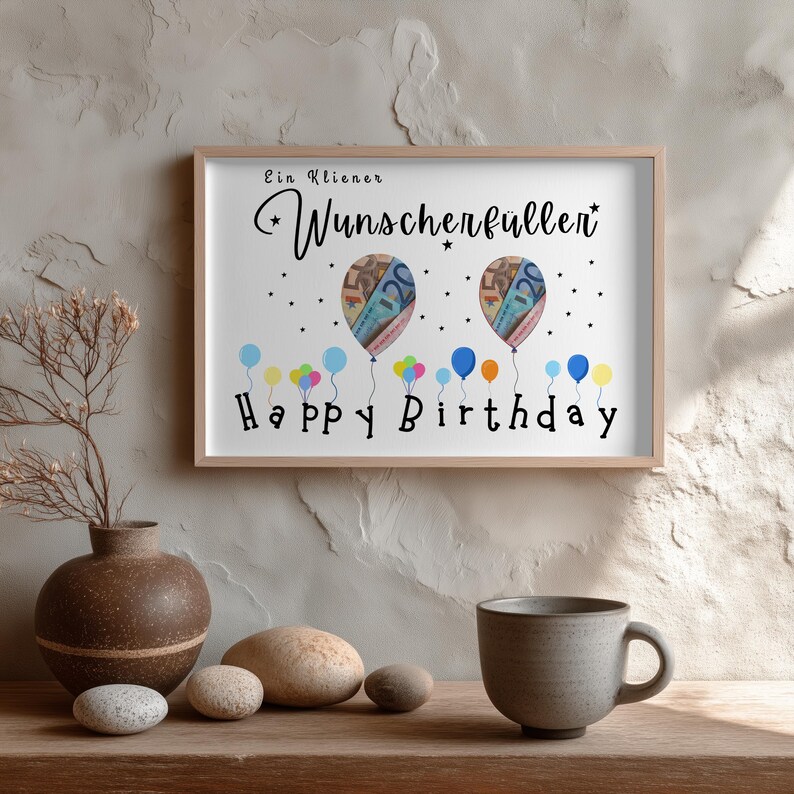 May include: A framed print with the words "Ein kleiner Wunscherf&uuml;ller" and "Happy Birthday". The artwork features two balloon illustrations with money designs, and colorful balloons at the bottom. The frame is light brown.