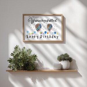 May include: A framed print with the words "Ein kleiner Wunscherf&uuml;ller" and "Happy Birthday". The artwork features two balloon illustrations with money inside and colorful balloons at the bottom. The frame is light brown.