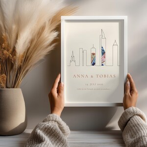 K&ouml;nnte beinhalten: Ein gerahmter Kunstdruck mit einem minimalistischen Skyline-Design. Der Druck enth&auml;lt die Namen "ANNA & TOBIAS" und das Datum "14. JULI 2026". Das Kunstwerk wird von zwei H&auml;nden gehalten, und der Druck hat einen cremefarbenen Hintergrund.