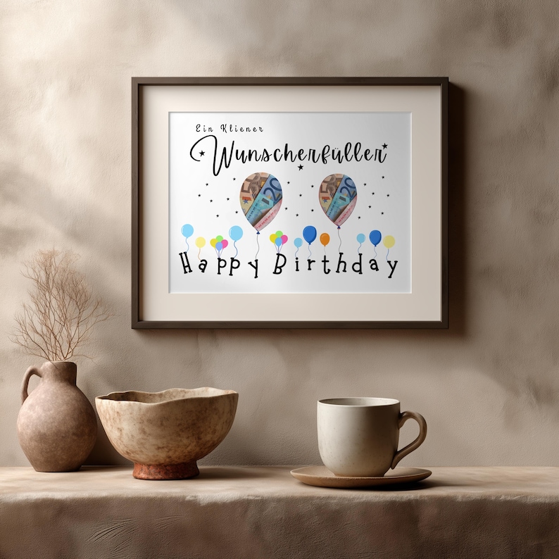 May include: A framed print with the words "Happy Birthday" and "Ein kleiner Wunscherf&uuml;ller" in black script. The artwork features two balloon illustrations with money inside, and colorful balloons at the bottom. The frame is dark brown.