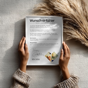 May include: A white framed print with German text, held by hands. The print includes the words "Wunsch-er-f&uuml;l-ler" and "Happy Birthday" with a small collage of money. The background is a neutral color.