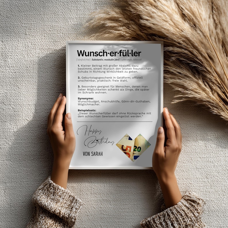 May include: A white framed print with German text, held by hands. The print includes the words "Wunsch-er-f&uuml;l-ler" and "Happy Birthday" with a small collage of money. The background is a neutral color.