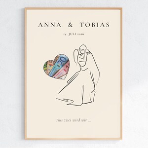 May include: A framed print with a beige background features the names "ANNA & TOBIAS" and the date "14. JULI 2026". A heart-shaped cutout reveals colorful currency. A line drawing depicts a couple embracing. The text "Aus zwei wird wir ..." is at the bottom.