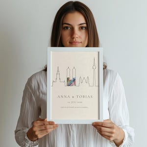 K&ouml;nnte beinhalten: Ein wei&szlig; gerahmter Druck mit einer minimalistischen Strichzeichnung einer Stadtsilhouette. Das Kunstwerk enth&auml;lt die Namen "ANNA & TOBIAS" und das Datum "14. JULI 2026".