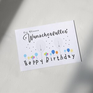 May include: White birthday card with the text "Ein kleiner Wunscherfullen" and "Happy Birthday". The card features illustrations of balloons in various colors, including blue, yellow, and pink.