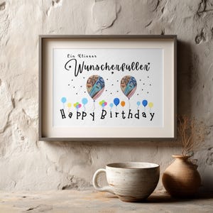 May include: A framed print with the words "Ein Kleiner Wunscherf&uuml;ller" and "Happy Birthday". The artwork features two balloons with money designs and colorful balloons at the bottom. A ceramic mug and vase are in the foreground.