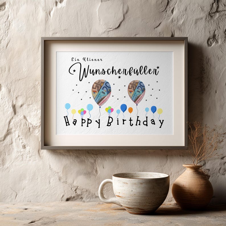 May include: A framed print with the words "Ein Kleiner Wunscherf&uuml;ller" and "Happy Birthday". The artwork features two balloons with money designs and colorful balloons at the bottom. A ceramic mug and vase are in the foreground.