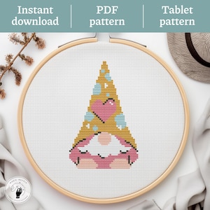 May include: A cross-stitch pattern of a gnome with a yellow hat, pink heart, and blue accents, set in a wooden embroidery hoop. The words "Instant download", "PDF pattern", and "Tablet pattern" are displayed above the hoop.