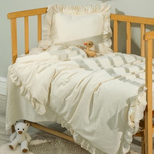 Puede incluir: Una cuna color crema con un edredón y una almohada a juego con volantes. Un pequeño pato de peluche y un oso de peluche están en la cama. La cuna es de madera clara, y la pared de atrás tiene obras de arte infantiles enmarcadas.