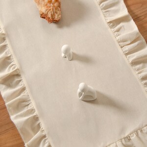 May include: Cream-colored table runner with ruffled edges. Two small white ceramic figurines and a piece of rough, tan-colored rock are placed on the runner. The runner is on a wooden surface.