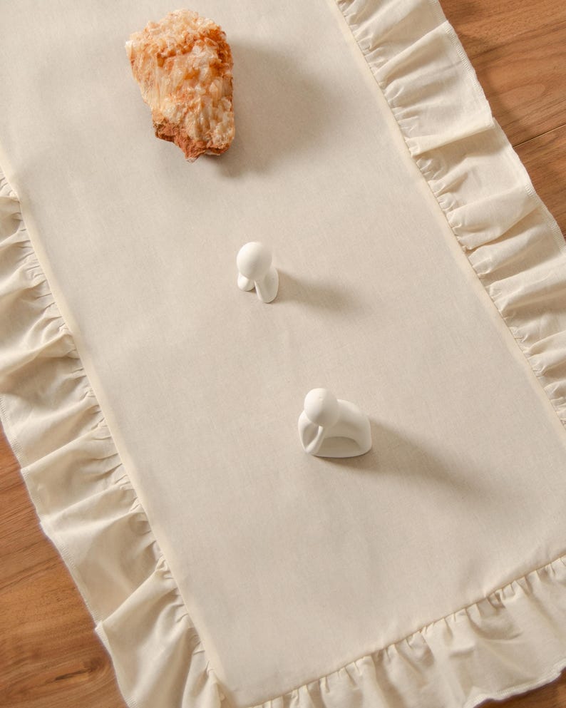 May include: Cream-colored table runner with ruffled edges. Two small white ceramic figurines and a piece of rough, tan-colored rock are placed on the runner. The runner is on a wooden surface.