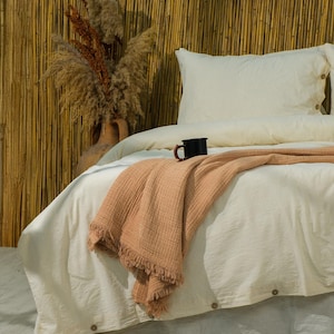 Può includere: Un letto con biancheria da letto color crema, una coperta con frange color pesca e una tazza nera. La testiera è una parete di bambù. Un vaso con piante secche si trova a sinistra del letto. La biancheria da letto ha bottoni.