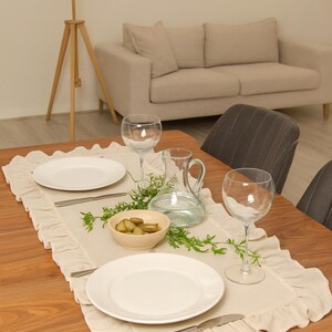 May include: A dining table set for a meal, featuring a cream-colored ruffled table runner. White plates, silverware, wine glasses, a water pitcher, and a bowl of pickles are arranged on the table. The table is made of wood.