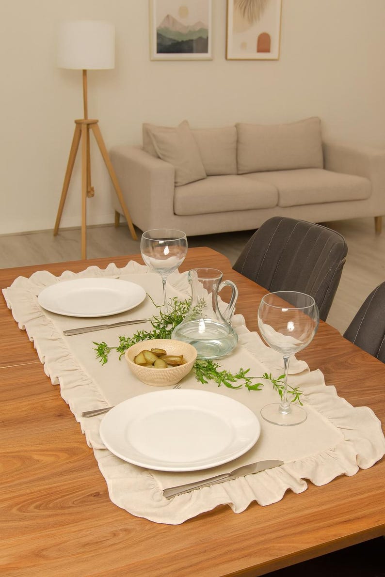 May include: A dining table set for a meal, featuring a cream-colored ruffled table runner. White plates, silverware, wine glasses, a water pitcher, and a bowl of pickles are arranged on the table. The table is made of wood.