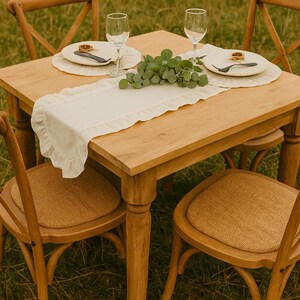 May include: A wooden table set for a meal, with four chairs. The table is set with plates, silverware, and wine glasses. A white runner with a ruffled edge runs down the center of the table. The chairs have woven seats.