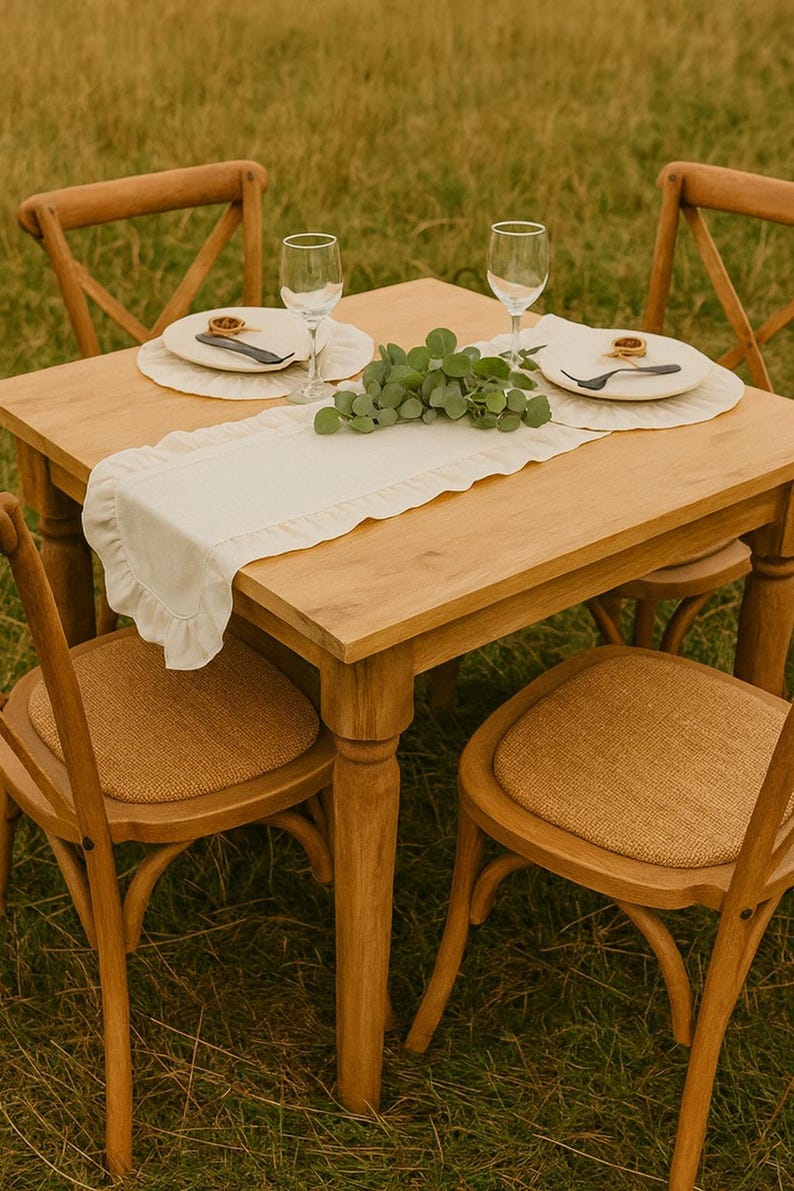 May include: A wooden table set for a meal, with four chairs. The table is set with plates, silverware, and wine glasses. A white runner with a ruffled edge runs down the center of the table. The chairs have woven seats.