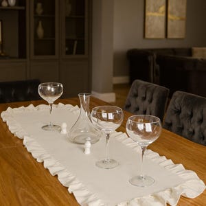 May include: A dining table set for a formal occasion. The table is adorned with a white ruffled runner, clear wine glasses, a decanter, and small white stoppers. The room has a warm, inviting ambiance.