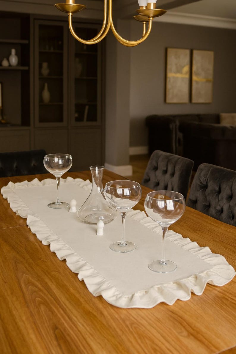 May include: A dining table set for a formal occasion. The table is adorned with a white ruffled runner, clear wine glasses, a decanter, and small white stoppers. The room has a warm, inviting ambiance.