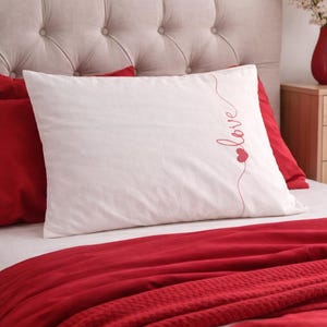 May include: A white pillow with the word "love" embroidered in red script and a heart design. The pillow is on a bed with red bedding and a red blanket. The headboard is beige with button tufting.