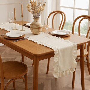 May include: A wooden dining table set with a cream-colored ruffled table runner, white plates, and champagne flutes. The table is set for a meal, with a vase of dried flowers and candles.