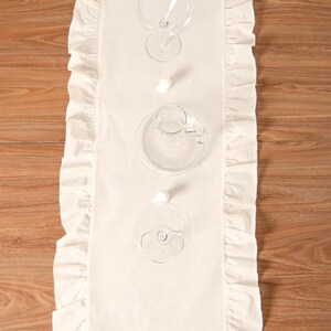 May include: Cream-colored rectangular table runner with ruffled edges, set with clear glassware. The runner is placed on a wooden surface. The glassware includes wine glasses, a small pitcher, and a saucer.