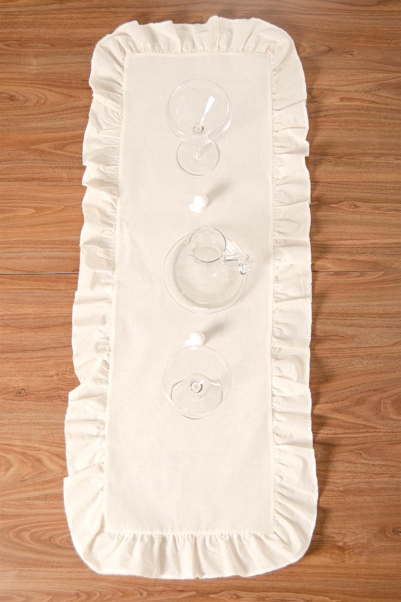 May include: Cream-colored rectangular table runner with ruffled edges, set with clear glassware. The runner is placed on a wooden surface. The glassware includes wine glasses, a small pitcher, and a saucer.