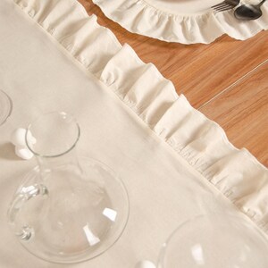 May include: Cream-colored linen table runner with ruffled edges, alongside a matching placemat and plate. The table setting includes a clear glass carafe, wine glasses, and silverware. Dried citrus slices adorn the plate.