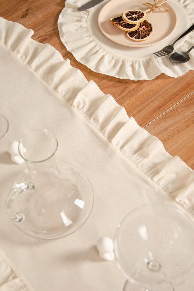May include: Cream-colored linen table runner with ruffled edges, alongside a matching placemat and plate. The table setting includes a clear glass carafe, wine glasses, and silverware. Dried citrus slices adorn the plate.
